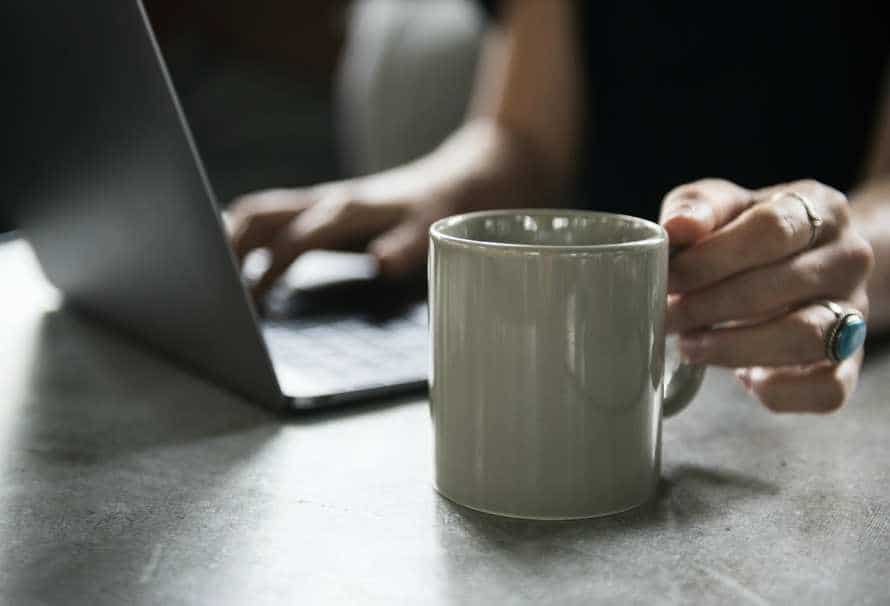 A person sitting at a table with a laptop and a coffee mug, enjoying a beverage while working.