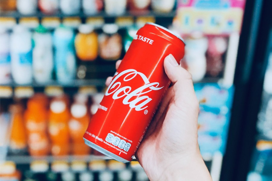 An inviting scene at a coffee shop in San Marcos, TX: A hand holding a refreshing can of Coke amidst a vibrant display of assorted beverages in a cooler.