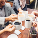 A group of people sitting at a table together doing a toast with coffee mugs.