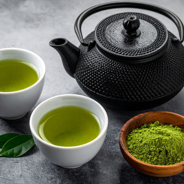 A black teapot sits beside two white cups filled with green tea, along with a small wooden bowl containing matcha powder, all arranged on a textured gray surface.