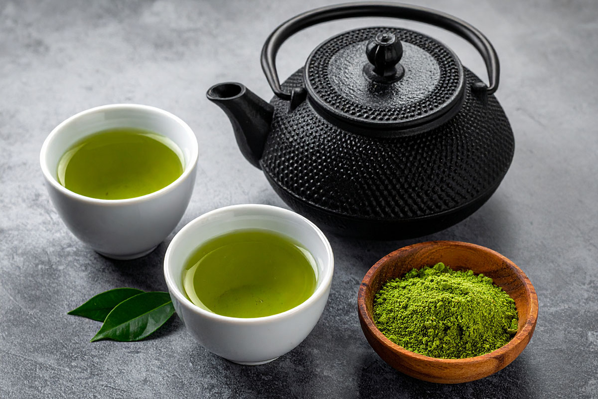 A black teapot sits beside two white cups filled with green tea, along with a small wooden bowl containing matcha powder, all arranged on a textured gray surface.