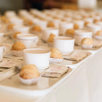 Rows of prepared trays with coffee cups and croissants line a counter at an event.