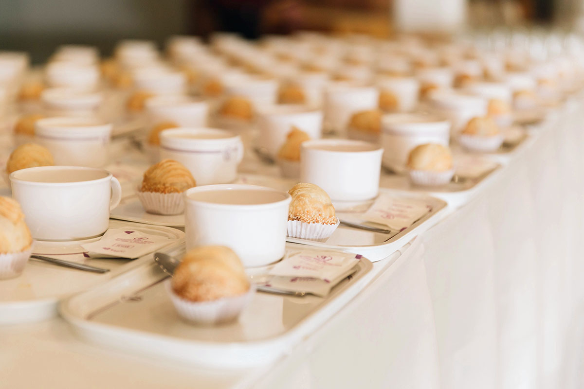 Rows of prepared trays with coffee cups and croissants line a counter at an event.