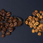 Two ornate silver spoons hold coffee beans: one spoon contains dark roasted beans, while the other features light roasted beans, both set against a dark background.
