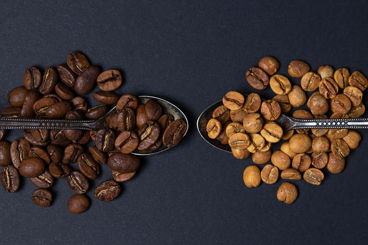 Two ornate silver spoons hold coffee beans: one spoon contains dark roasted beans, while the other features light roasted beans, both set against a dark background.