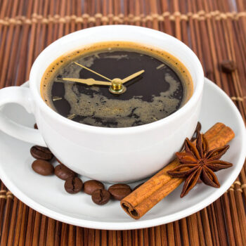 A white coffee cup with a frothy surface designs resembling a clock, surrounded by coffee beans and star anise on a woven brown mat.