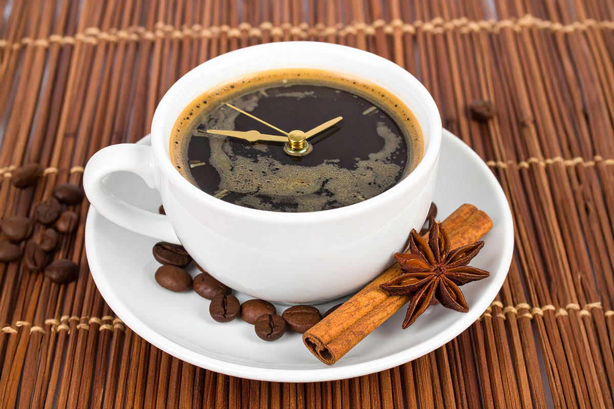 A white coffee cup with a frothy surface designs resembling a clock, surrounded by coffee beans and star anise on a woven brown mat.