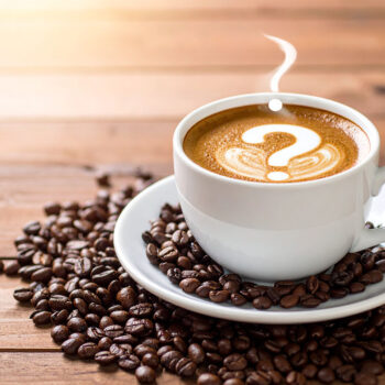 A white cup of coffee on a matching saucer, with a question mark pattern formed by the foam on the surface, resting on a wooden table.