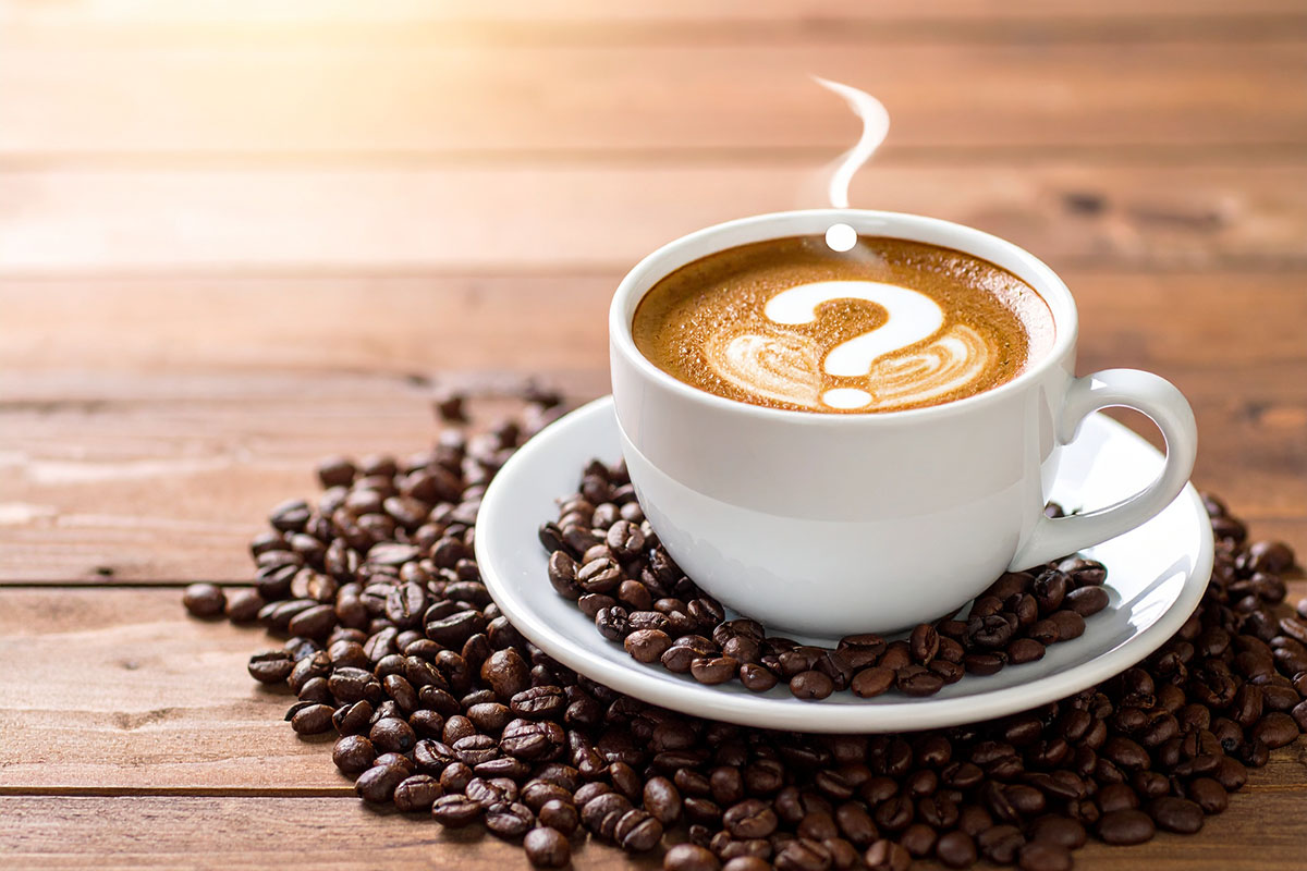 A white cup of coffee on a matching saucer, with a question mark pattern formed by the foam on the surface, resting on a wooden table.
