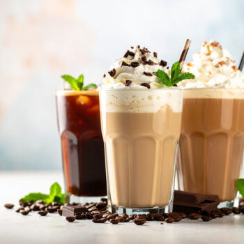 Three elegantly presented coffee beverages are displayed: the front cup contains a creamy coffee topped with whipped cream, chocolate shavings, and a mint leaf; the back left features a dark iced coffee with a straw; and the back right showcases a layered drink with whipped cream. A light background enhances the vibrant colors of the drinks.