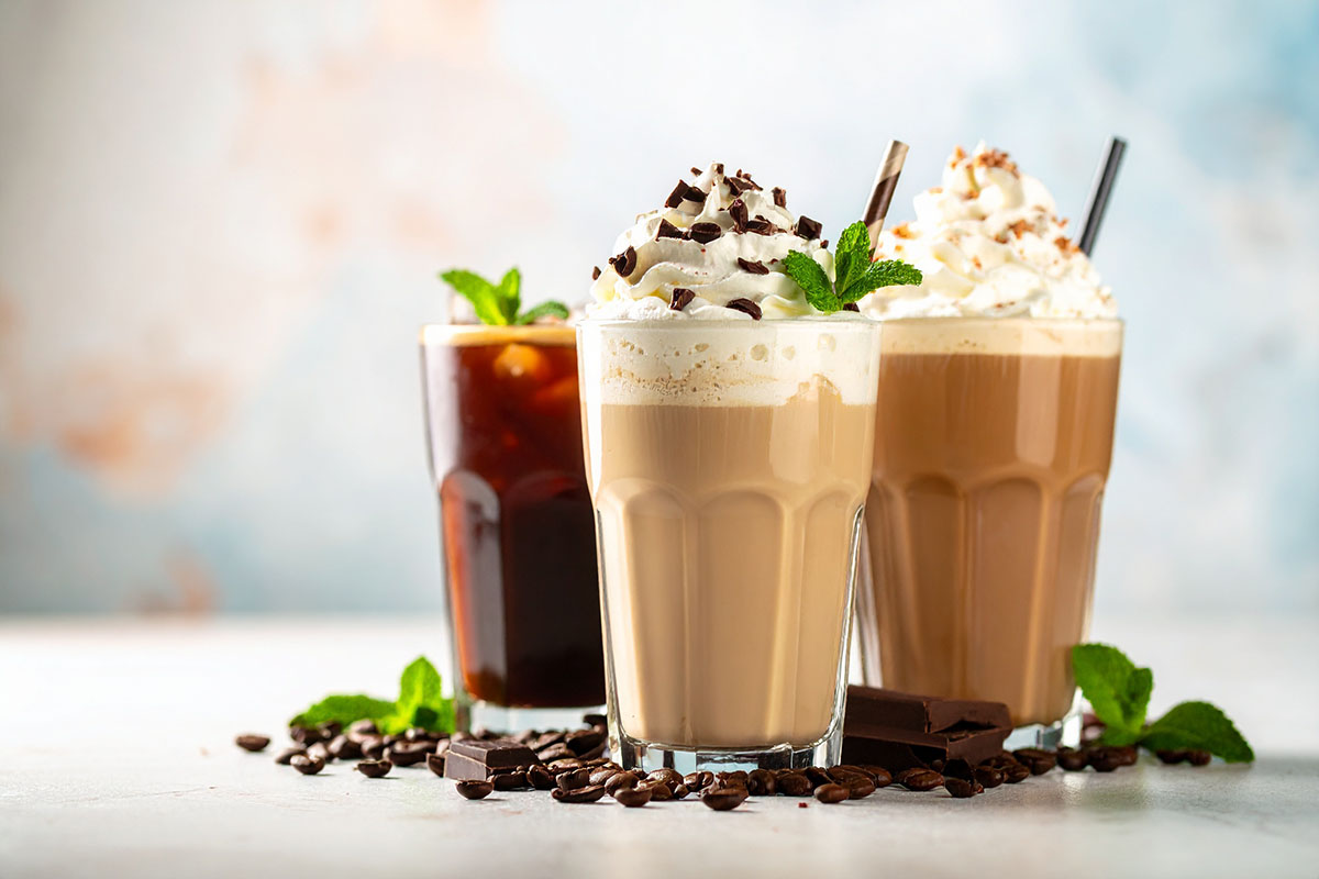 Three elegantly presented coffee beverages are displayed: the front cup contains a creamy coffee topped with whipped cream, chocolate shavings, and a mint leaf; the back left features a dark iced coffee with a straw; and the back right showcases a layered drink with whipped cream. A light background enhances the vibrant colors of the drinks.