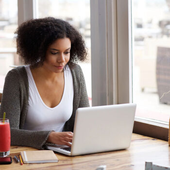 A young woman with curly hair sits at a wooden table in a cafe, focused on her laptop. She has a pink smoothie beside her and a notebook with pens in front of her, while colorful flowers are visible in the background near the window.