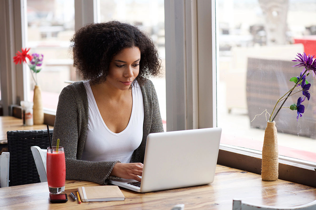 A young woman with curly hair sits at a wooden table in a cafe, focused on her laptop. She has a pink smoothie beside her and a notebook with pens in front of her, while colorful flowers are visible in the background near the window.
