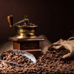 A vintage coffee grinder beside a wooden box, with whole coffee beans scattered around and a metal scoop filled with beans resting on the table. The background is softly lit, enhancing the warm tones of the coffee.