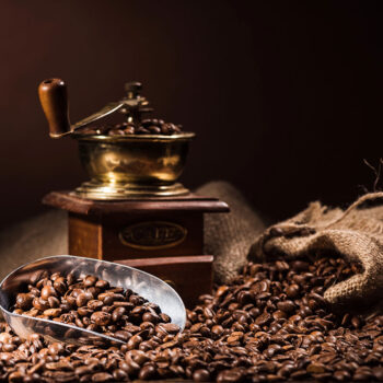 A vintage coffee grinder beside a wooden box, with whole coffee beans scattered around and a metal scoop filled with beans resting on the table. The background is softly lit, enhancing the warm tones of the coffee.