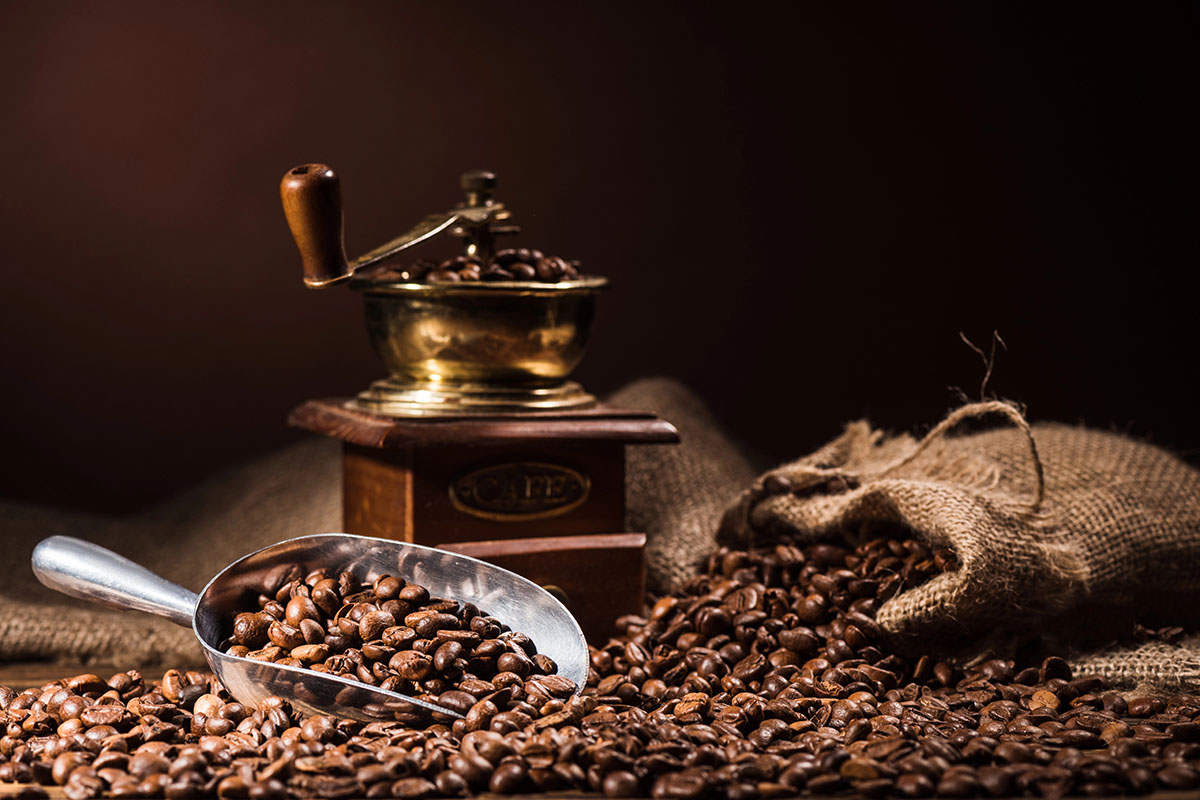 A vintage coffee grinder beside a wooden box, with whole coffee beans scattered around and a metal scoop filled with beans resting on the table. The background is softly lit, enhancing the warm tones of the coffee.