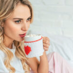 A woman with long, wavy hair sits on a bed, holding a mug decorated with a red heart. She gazes thoughtfully at the mug filled with hot chocolate and marshmallows. The background features a light-colored brick wall and soft bedding.