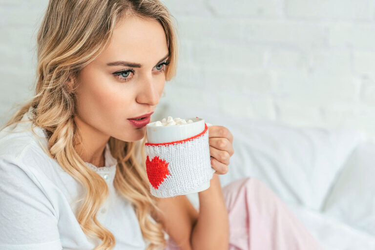 A woman with long, wavy hair sits on a bed, holding a mug decorated with a red heart. She gazes thoughtfully at the mug filled with hot chocolate and marshmallows. The background features a light-colored brick wall and soft bedding.
