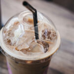 A close-up of a plastic cup containing iced coffee, with ice cubes visible and a black straw inserted through the clear lid, set on a wooden table.