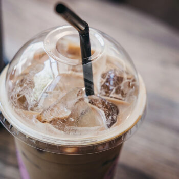A close-up of a plastic cup containing iced coffee, with ice cubes visible and a black straw inserted through the clear lid, set on a wooden table.