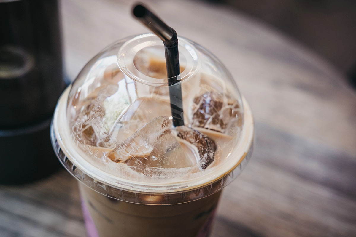 A close-up of a plastic cup containing iced coffee, with ice cubes visible and a black straw inserted through the clear lid, set on a wooden table.