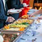 A buffet table with a variety of dishes, including rice, vegetables, and appetizers, as individuals in formal attire serve themselves food onto their plates.