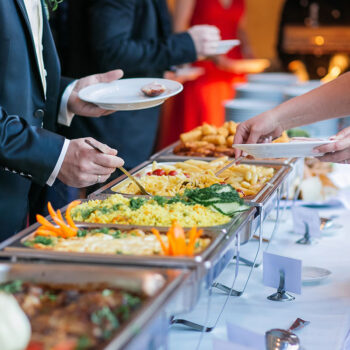 A buffet table with a variety of dishes, including rice, vegetables, and appetizers, as individuals in formal attire serve themselves food onto their plates.
