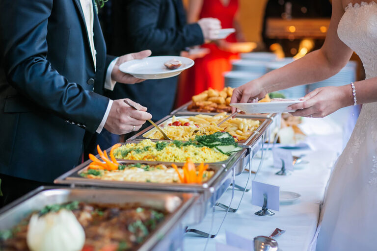 A buffet table with a variety of dishes, including rice, vegetables, and appetizers, as individuals in formal attire serve themselves food onto their plates.