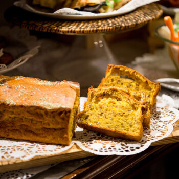 A loaf of golden-brown bread with a slice cut out, revealing its crumb texture, sits on a lace doily atop a wooden board. In the background, there's a selection of appetizers and a bowl of colorful vegetables.