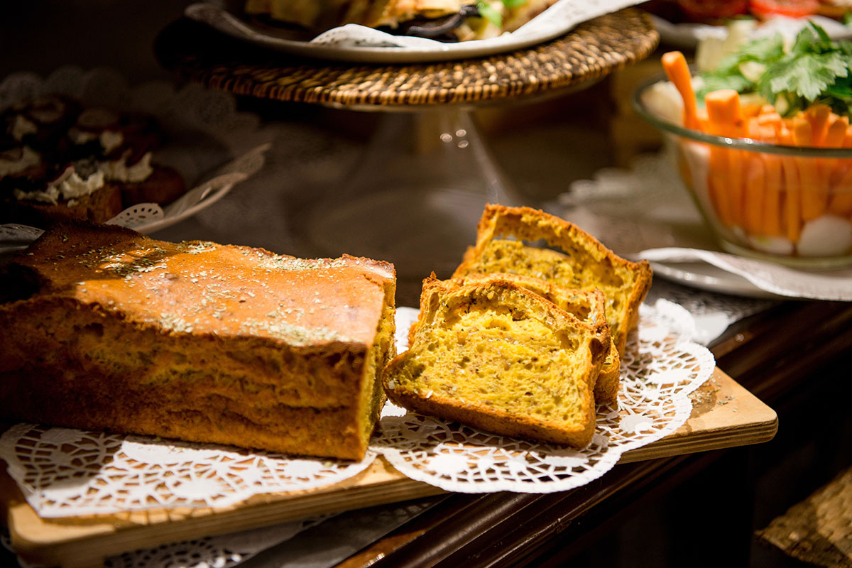 A loaf of golden-brown bread with a slice cut out, revealing its crumb texture, sits on a lace doily atop a wooden board. In the background, there's a selection of appetizers and a bowl of colorful vegetables.