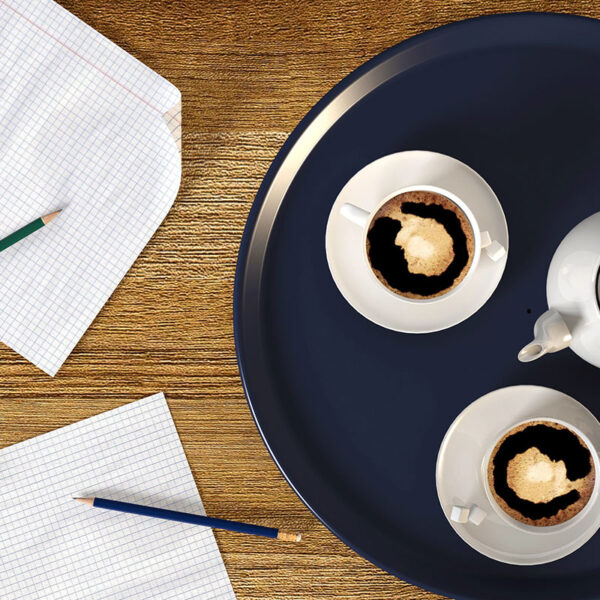 A wooden table with a dark round tray featuring two white cups of coffee and a teapot, accompanied by two sheets of graph paper and colored pencils.
