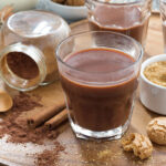 A wooden serving tray holds a glass of thick hot chocolate, surrounded by cinnamon sticks, a jar of cocoa powder, a small bowl of brown sugar, and broken pieces of cookies.