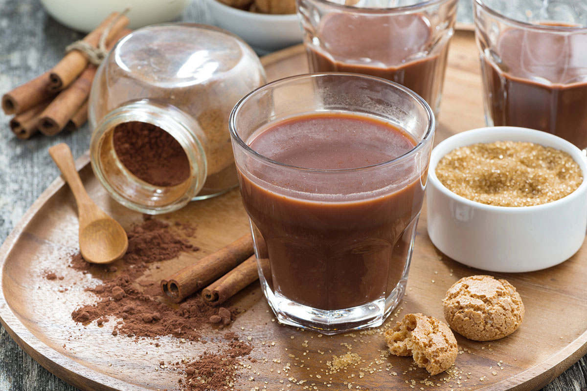 A wooden serving tray holds a glass of thick hot chocolate, surrounded by cinnamon sticks, a jar of cocoa powder, a small bowl of brown sugar, and broken pieces of cookies.