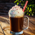 A glass of chocolate beverage topped with whipped cream and crushed peppermint, featuring a red and white striped straw, set on a wooden table with a hint of greenery in the background.
