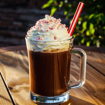 A glass of chocolate beverage topped with whipped cream and crushed peppermint, featuring a red and white striped straw, set on a wooden table with a hint of greenery in the background.
