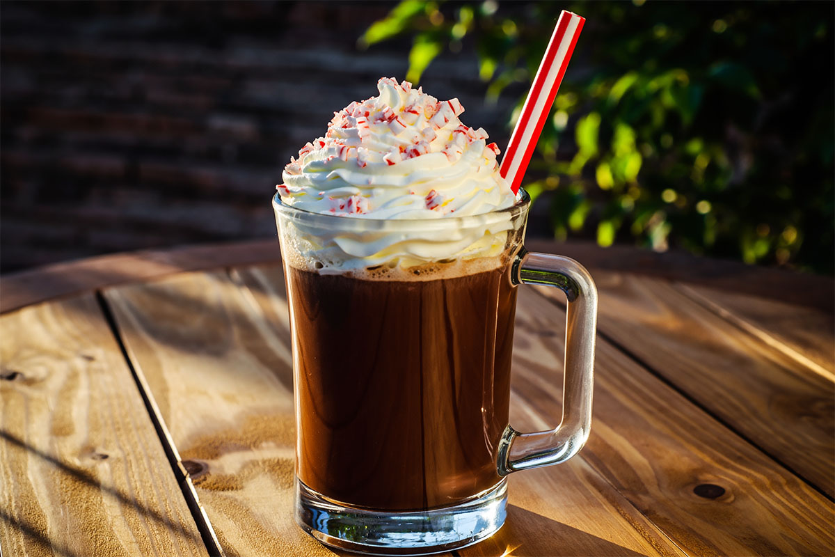 A glass of chocolate beverage topped with whipped cream and crushed peppermint, featuring a red and white striped straw, set on a wooden table with a hint of greenery in the background.