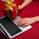 A hand placing a red gift box with a gold ribbon on a laptop, set against a red background.