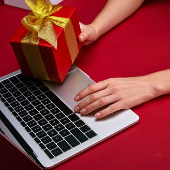 A hand placing a red gift box with a gold ribbon on a laptop, set against a red background.