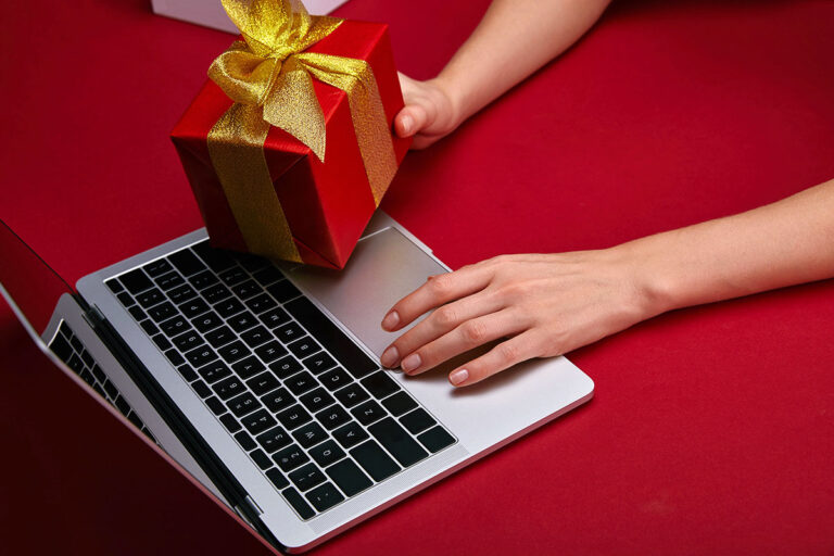 A hand placing a red gift box with a gold ribbon on a laptop, set against a red background.