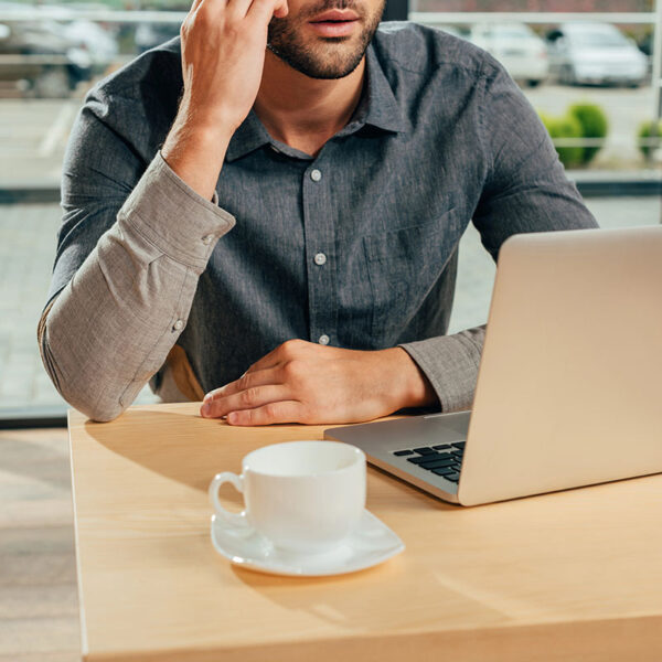 A man in a gray button-up shirt sits at a wooden table, resting his chin on his hand while looking at a laptop. A small white coffee cup is on a saucer in front of him. The background features large windows with a view of parked cars and greenery.