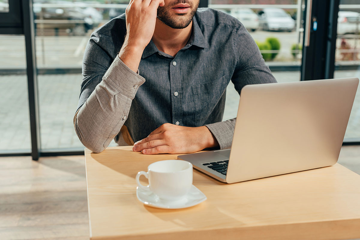 A man in a gray button-up shirt sits at a wooden table, resting his chin on his hand while looking at a laptop. A small white coffee cup is on a saucer in front of him. The background features large windows with a view of parked cars and greenery.