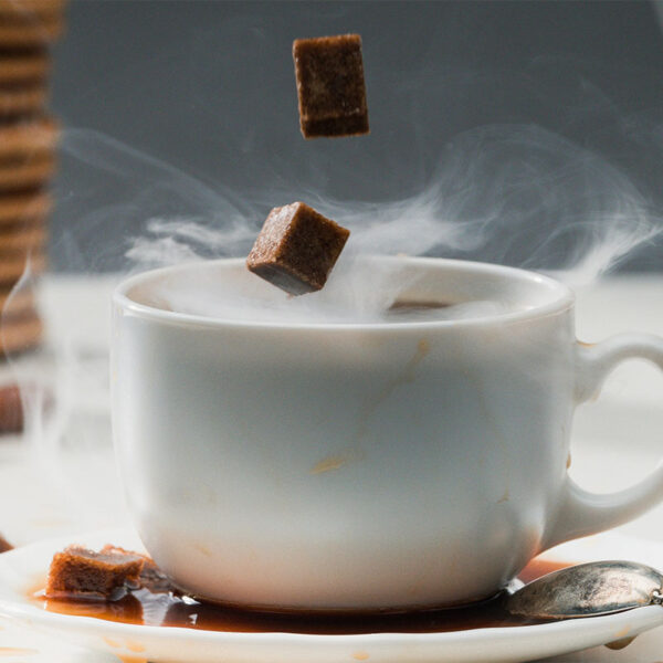 A steaming cup of coffee with two sugar cubes suspended above it, resting on a saucer with a silver spoon, surrounded by scattered spices and a stack of cookies in the background.