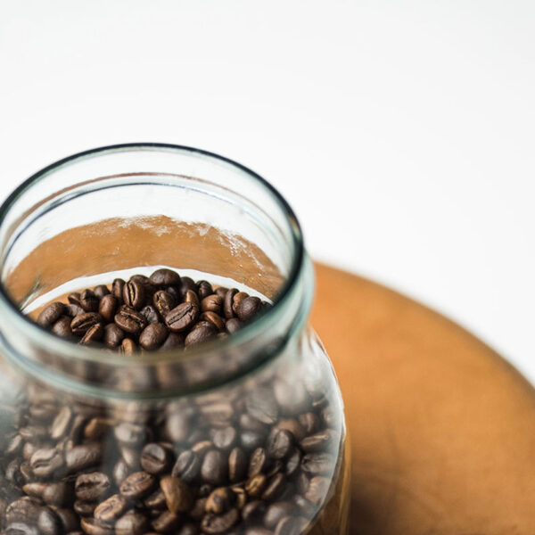 A glass jar filled with coffee beans sits on a wooden surface against a white background.