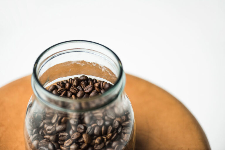A glass jar filled with coffee beans sits on a wooden surface against a white background.