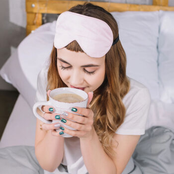 A woman wearing a pink sleep mask is sitting on a bed, holding a large cup of coffee close to her face, with a content expression. Her nails are painted teal with one accent nail featuring glitter. The background contains white bedding and a wooden headboard.