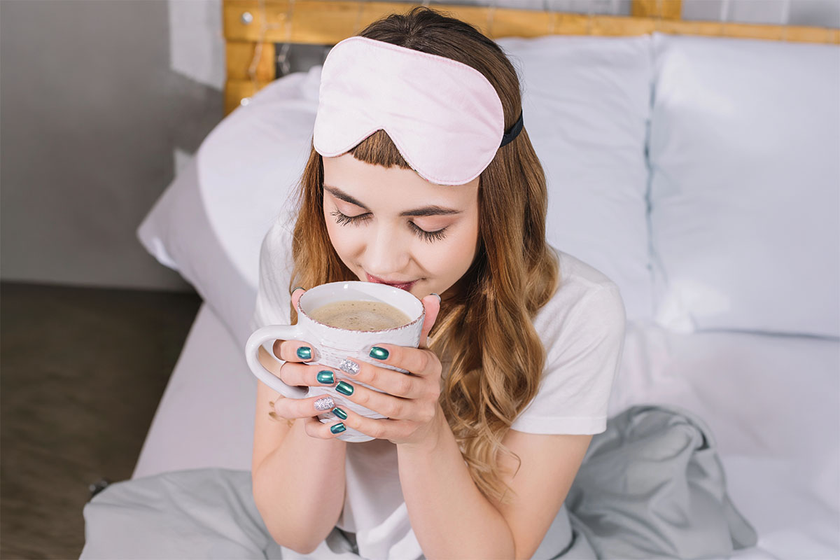 A woman wearing a pink sleep mask is sitting on a bed, holding a large cup of coffee close to her face, with a content expression. Her nails are painted teal with one accent nail featuring glitter. The background contains white bedding and a wooden headboard.