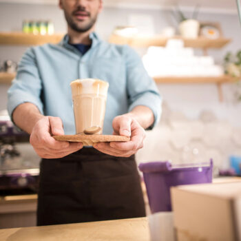 A barista in a light blue shirt serves a latte in a glass, placed on a wooden tray, from behind a coffee counter with various items visible in the background.