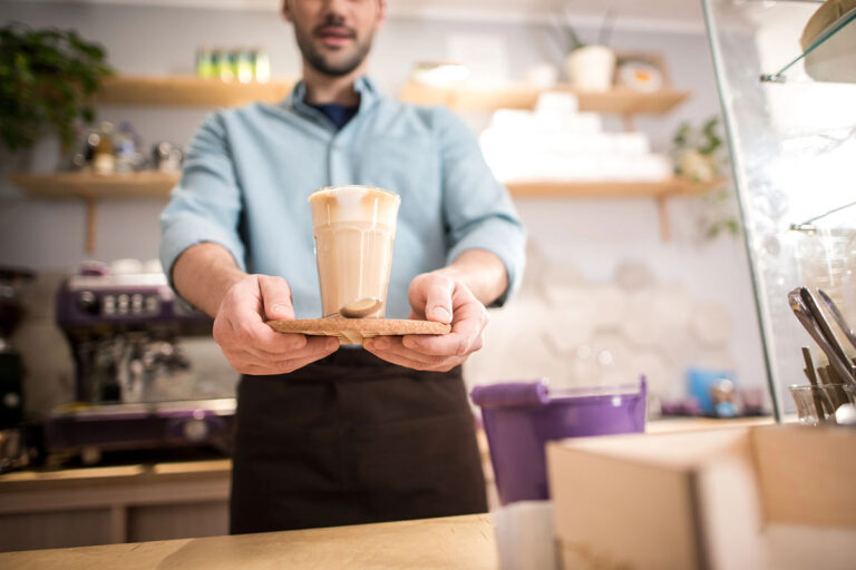 A barista in a light blue shirt serves a latte in a glass, placed on a wooden tray, from behind a coffee counter with various items visible in the background.
