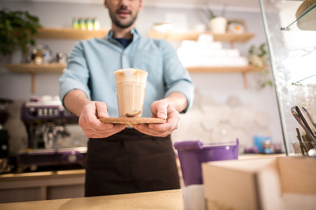 A barista in a light blue shirt serves a latte in a glass, placed on a wooden tray, from behind a coffee counter with various items visible in the background.