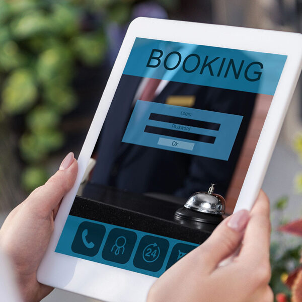 A person holding a tablet displaying an online booking interface with fields for login and password, alongside a service bell on a counter and greenery in the background.